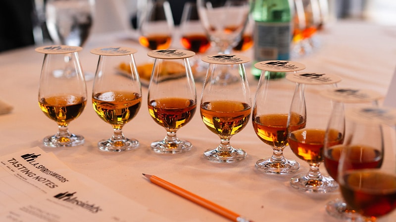 Tasting glasses from the LA Spirits awards competiton lined up on a table in a curve shape with coasters covering the liquid and a pencil on the table under the curve of glasses.